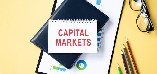 Handwriting text writing Capital Markets. Concept meaning buyers and sellers engage in trade of financial securities Paper placed tilted above buffer wire on soft pastel multi colours backdrop.