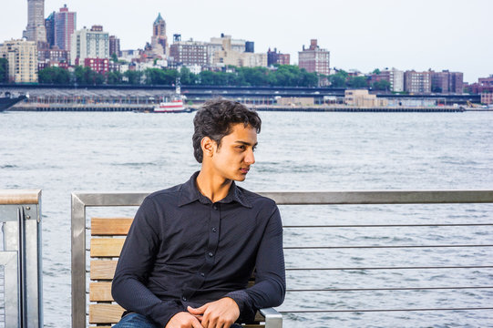 18 Years Old East Indian American Male Teenager Relaxing Outside In New York City, Wearing Black Long Sleeve Shirt, Sitting On Chair By East River, Looking Around, Thinking..