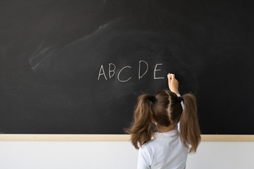 Sweet little girl at school in a lesson. A child stands in front of a black board. She writes in English the letters of the alphabet in chalk. Place for an inscription. Education concept.