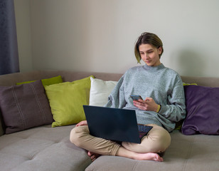 serious teenage girl blogger working online on laptop with smartphone on sofa at home. Concept of distance education or learning, online shopping, communication with friends