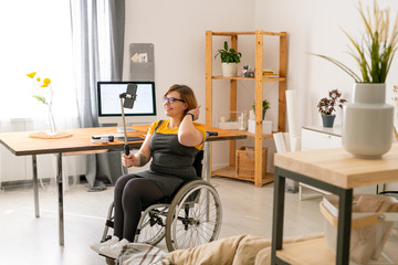 Fototapeta premium Cheerful disabled young female blogger in glasses sitting in wheelchair and streaming video at home