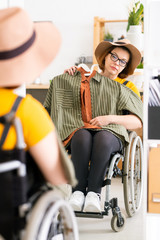 Attractive young woman in hat and eyeglasses sitting in wheelchair and choosing outfit in front of mirror