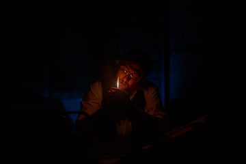 A young man in a white shirt with glasses sits and wants to set fire to a lighter. Photo in the twilight. Selective focus. home work, freelance, online learning, studying concept.