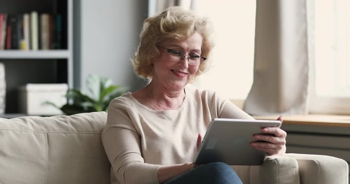 Happy attractive middle aged blonde retired woman in eyeglasses using digital computer tablet, relaxing on comfortable sofa alone at home. Smiling older lady enjoying shopping, communicating online.