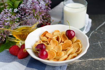 Tiny pancake cereal, cherry, strawberry, honey and milk on dark rustic background with lilac flowers