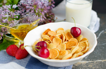 Tiny pancake cereal, cherry, strawberry, honey and milk on dark rustic background with lilac flowers