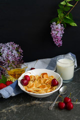 Tiny pancake cereal, cherry, strawberry, honey and milk on dark rustic background with lilac flowers