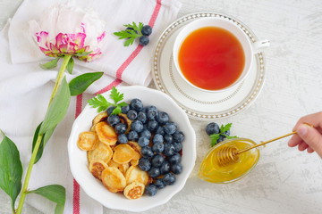 Tiny pancake cereal with blueberry, honey and tea for breakfast in the morning light and pink peony on table