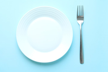A flat white empty plate and a metal fork on a blue background. Serving. Selective focus. Starvation