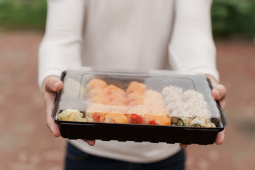 Closeup sushi in box healthy food delivery online service. Man in white clothes gives sushi set to you. Japanese cuisine: rolls, soy sauce, wasabi. Advert for restaurant