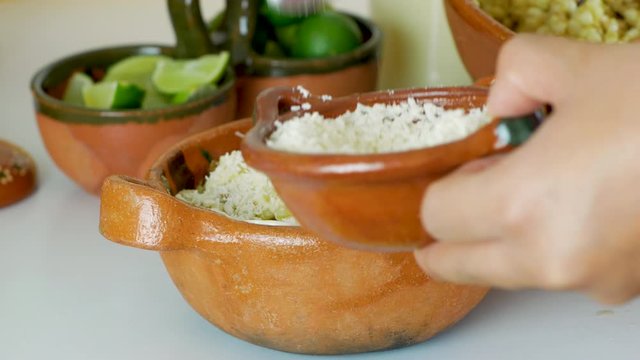 Woman hand's pours the grated cheese in Mexican Street Corn Salad (Esquites) into traditional clay dish