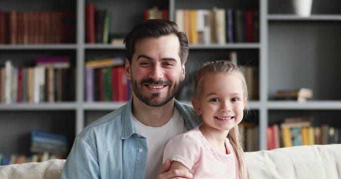 Close up head shot loving smiling handsome father holding on lap adorable small preschool kid daughter, sitting together on comfortable couch at home. Happy daddy looking at camera with little child.