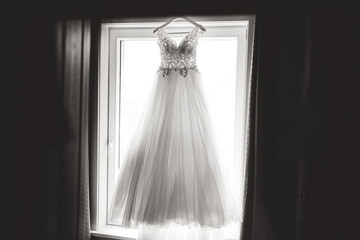 Black and white photo of lace wedding dress on hanger hanging on window frame and waiting for bride. Back-light photo. Wedding day concept.