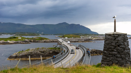 Cairn rock next to atlantic ocean road in Molde, Norway. Seagull sitting on top with mountains in the background. Tourists are fishing from the bridge with cars driving by.