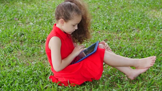 Curious cute girl preschool child using a digital tablet technology device looks telephone sitting on the street on the grass in the park. Kid plays in the tablet.