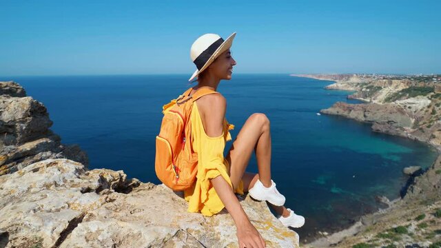 Happy atractive taned woman posing with hand reaching out on cliff edge with beautiful sea view. Carefree female in bright yellow dress and straw sunhat. Freedom, travel and vacation concept.