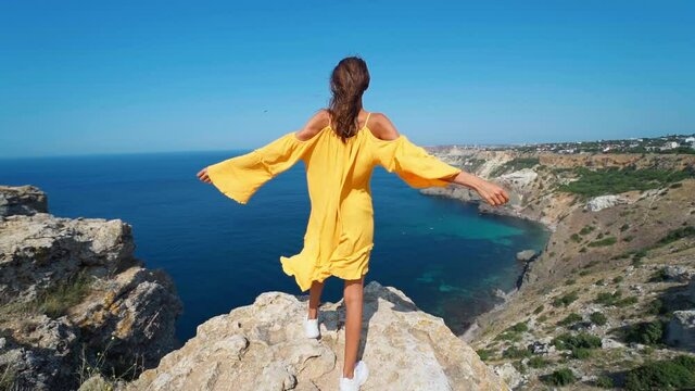 Full length epic slow motion of taned traveler woman standing on top rock cliff with open arms in front of amazing seascape, bright yellow dress and hair blowing in the wind.