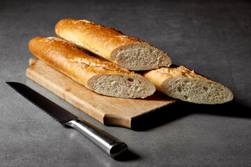 
Halved fresh baguette on a wooden board and a kitchen knife on a gray surface.