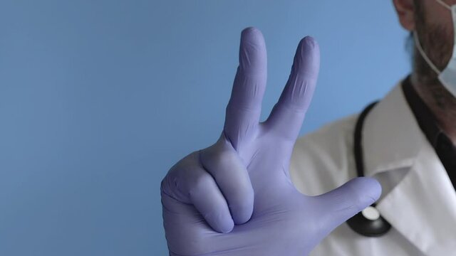 doctor in a white coat, gloves and stethoscope standing against the blue background and counting from 0 to 5 by the hand