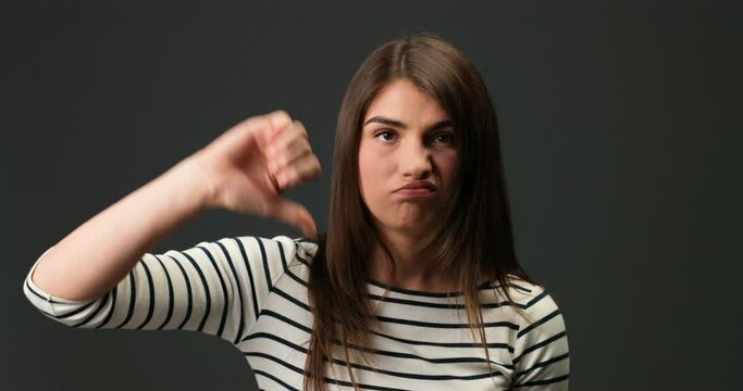 Close up portrait of unsatisfied beautiful Caucasian young female in striped blouse with funny face giving thumbs down. Unhappy pretty girl with dark hair over grey background. Emotions concept