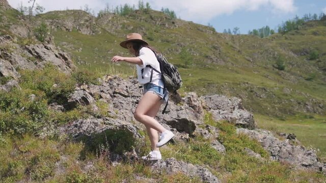 Attractive brunette girl in a hat with a backpack travels in the mountains. woman in a white shirt and shorts climbs the mountains