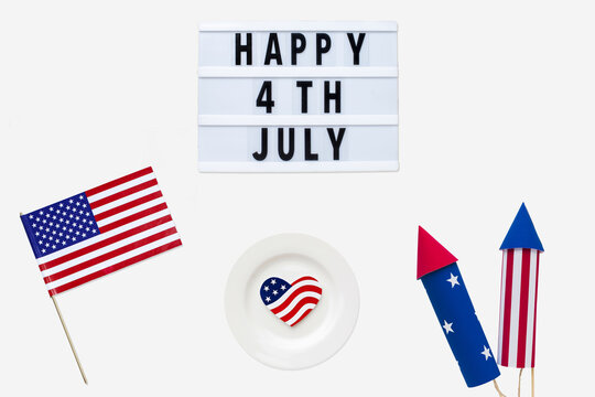 American flag, signboard with happy 4th july text, American flag shaped cookie and fireworks are placed in a white background. Flat lay with having space for text.