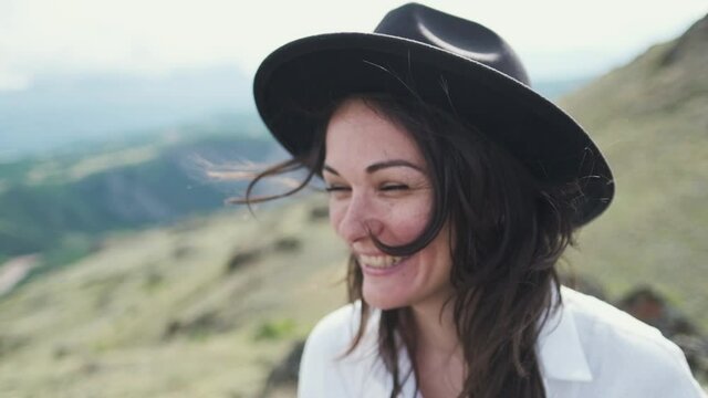 Attractive brunette in a hat and white shirt is enjoying nature in the mountains. hat and hair flying in the wind close-up. hand movement with sunbeams