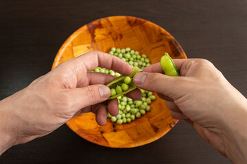 Closeup to a male hands threshing fresh green peas and a wooden bowl with green peas over a dark...