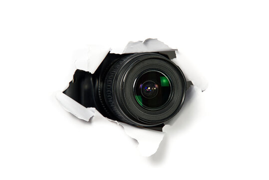 black camera with a telephoto lens that looks out through a hole in white paper. Concept of paparazzi, espionage, yellow press. camera lens looking through hole in wall.