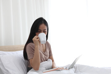 Woman relaxing and drinking cup of hot coffee or tea using laptop computer  in the bedroom.woman checking social apps and working.Communication and technology concept