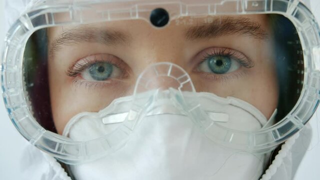 Close-up portrait of young girl doctor in safety suit goggles and respirator standing alone on white background and looking at camera. People and job concept.