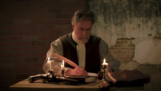 A candle lit desk where a flintlock pistol lays while an American 18th century colonist writes in his journal with a plumed quill pen.