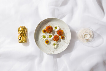 Top view of voile fabric background with a plate with floating flowers, a small golden buddha and a glass bowl with a candle and white rocks inside. Space for text.