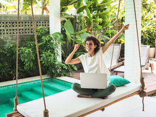 cheering digital nomad woman working with laptop on a bamboo hanging swing surrounded by green plants and pool in asia
