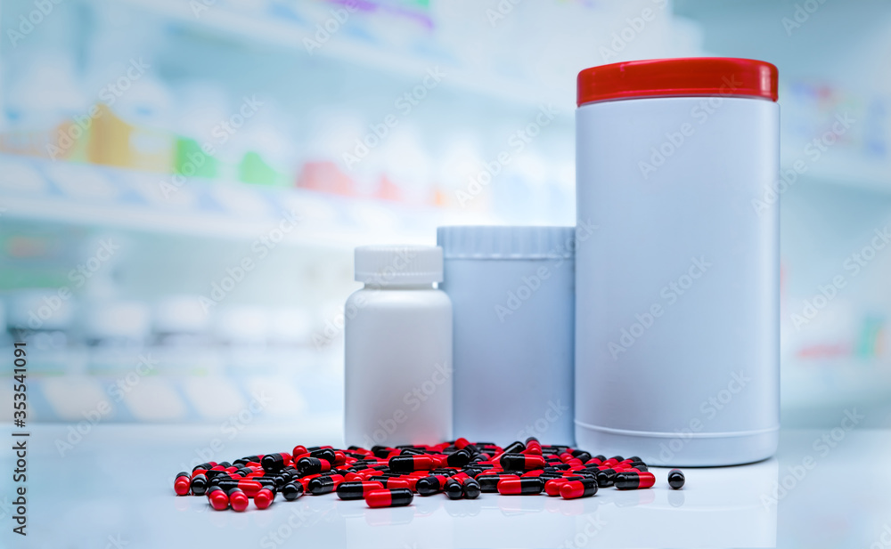 Wall mural Red-black capsules pills spread on counter in drugstore. Blank label plastic drug bottle on blurred background of vitamins and supplements display shelf in pharmacy shop. Prescription drug concept.