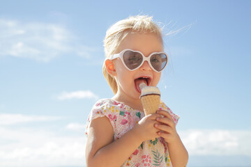 Adorable toddler girl eating ice cream. Portrait of child wearing sunglasses and holding an ice cream with beautiful blue sea and sky behind. Happy summer vacation on the beach.