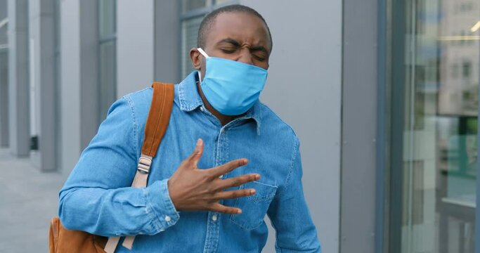African American young man in medical mask walking the street and feeling himself very bad. Coronavirus disease concept. Guy strolling outdoor, caughing and having pain in lungs.