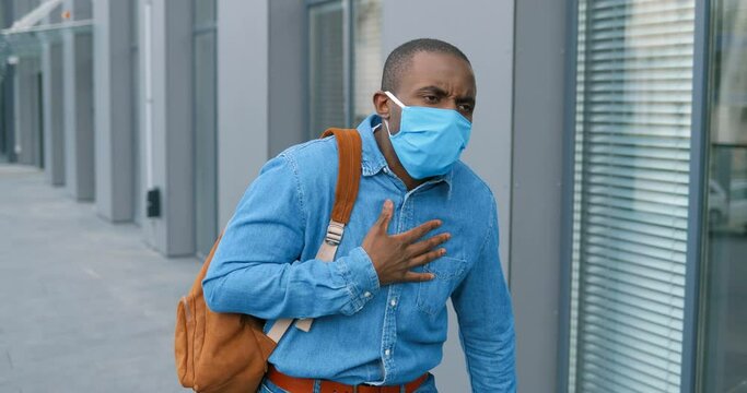 African American young man in medical mask walking the street and feeling himself very bad. Coronavirus disease concept. Guy strolling outdoor, caughing and having pain in lungs. Symptoms of covid-19.