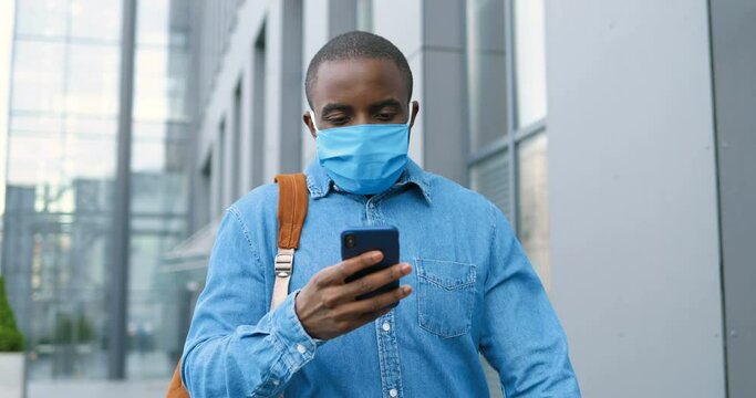 Male pedestrian in medical mask walking the street, looking for address and texting message on phone. African American man in respiratory protection strolling and tapping or scrolling on smartphone.