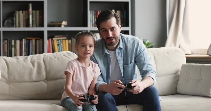 Happy young father teaching little preschool kid girl playing video games, resting together on comfortable sofa at home. Smiling daddy using joysticks, enjoying funny activity with child daughter.