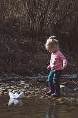 Little girl in rubber boots launches paper white boats in creek in spring or autumn