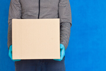 Courier hand holds a brown box on a blue background in rubber gloves on a face mask. Detail of a man carrying a cardboard parcel with copy space.