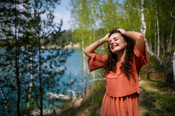Naklejka premium A girl with long wavy curly hair in her hair woven flowers in a white transparent guipure dress barefoot in the summer on nature in a forest at sunset standing standing near the road and bushes