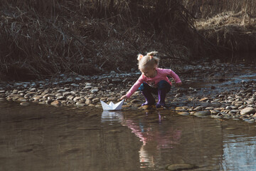 Little girl in rubber boots launches paper white boats in creek in spring or autumn