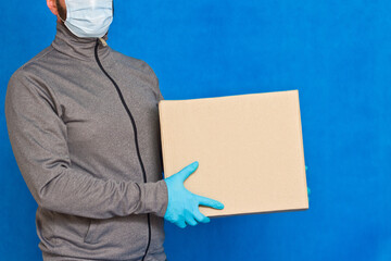 Courier hand holds a brown box on a blue background in rubber gloves on a face mask. Detail of a man carrying a cardboard parcel with copy space.