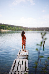 Fototapeta na wymiar Wooden masonry. A girl with long wavy curly hair in an orange guipure dress and shoes on nature, in a forest against a lake, stood near trees and bushes. Young woman smiles and enjoys life