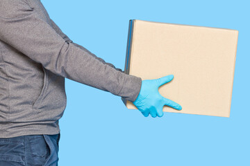 Courier hand holding a brown box on a blue background in rubber gloves on a face mask. Detail of a man carrying a cardboard parcel with copy space.