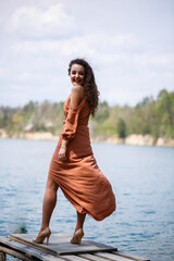 A young woman in a dress stands on wooden masonry in the middle of a blue lake. Girl happy smiles and the sun shines, summer day. She has curly long hair and a European appearance