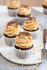 Peanut butter cupcakes with chocolate bites and chopped nuts on grey linen tablecloth.