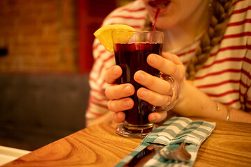 a glass cup with a delicious aromatic mulled wine is held in the hands of a girl in blur and drinks from a straw. soft focus, close up.